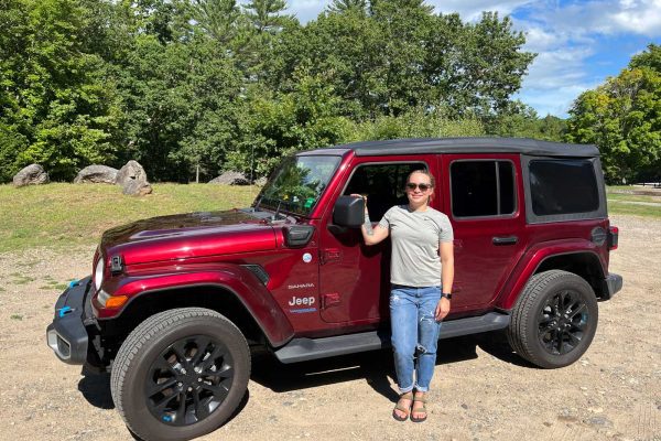 woman in front of red EV jeep in front of trees on a dirt road