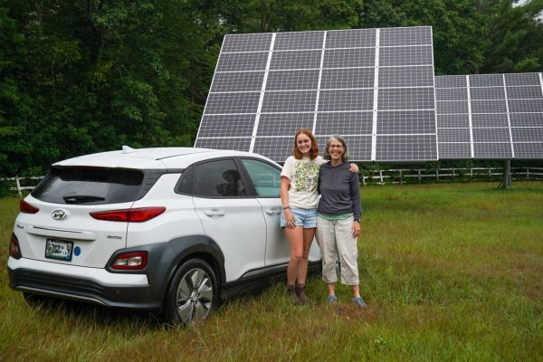 Two women in a field beside a white EV in front of solar panels