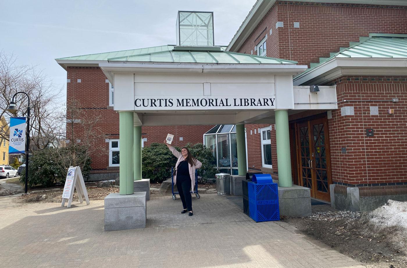 Woman stands in front of library holding a book in the air