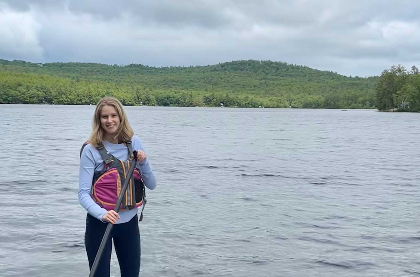 young woman smiling on paddleboard with mountains and pond behind her