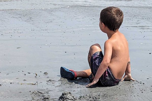 young boy sitting in sand on beach looking out at ocean