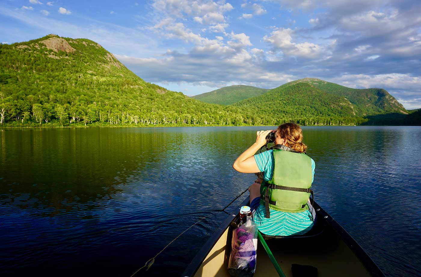 woman paddling on lake in a canoe, with mountains and blue sky ahead of her