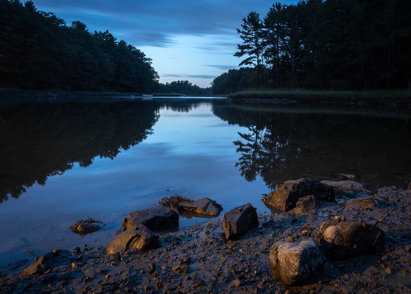 Twilight pic of water with trees on either side and rocks in foreground