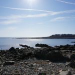 View of ocean under bright sun and blue sky, with rocky coast in foreground