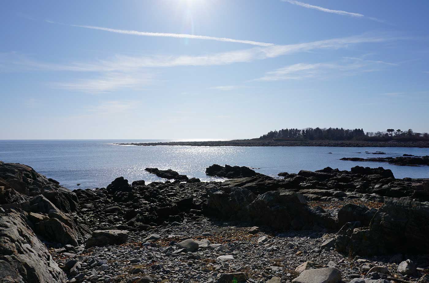 View of ocean under bright sun and blue sky, with rocky coast in foreground