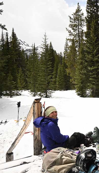 person in winter clothes sitting in snow, with evergreen trees behind them