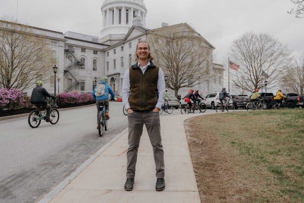 Josh Caldwell standing in front of State House with people riding electric bikes behind him