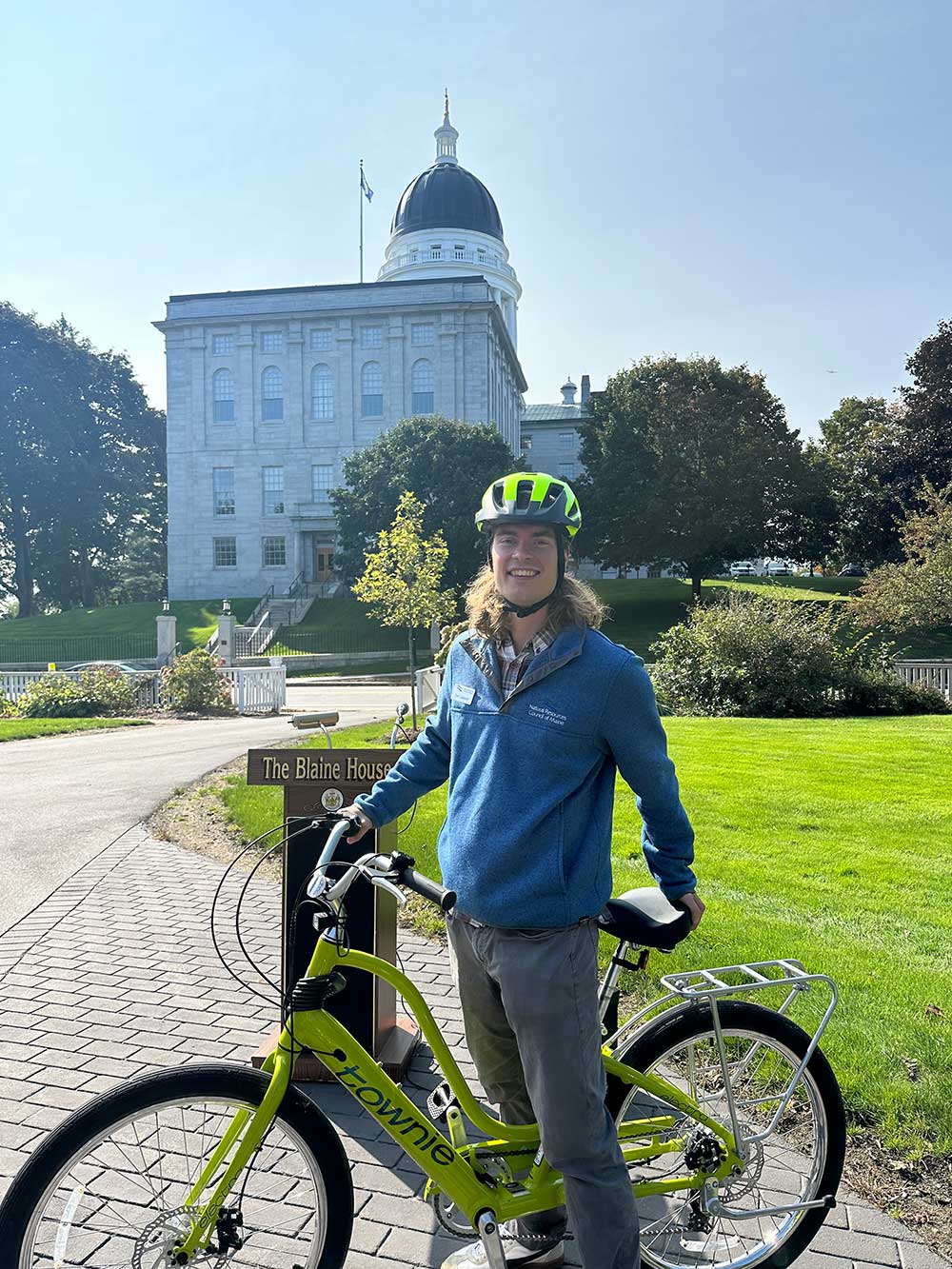person wearing helmet posing with e-bike in front of Maine State House