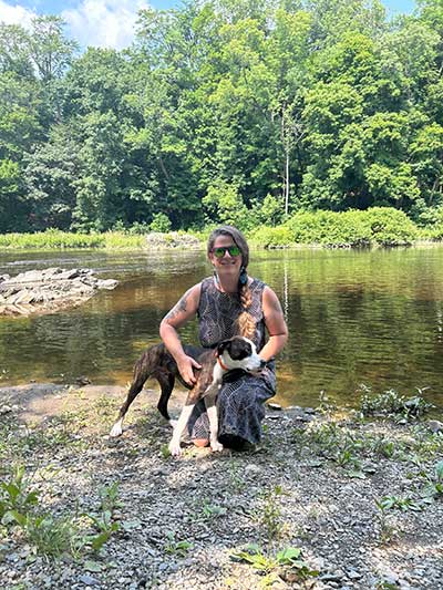 Mareisa holding dog while kneeling down in front of water and forests