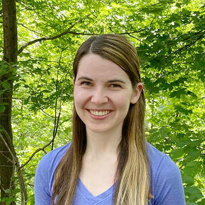 kate-phillips Young woman with long hair and purple shirt, smiling in front of trees
