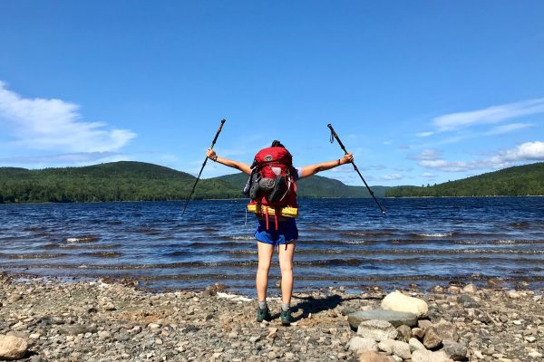 Person carrying backpack facing water and mountains holding hiking poles over their head