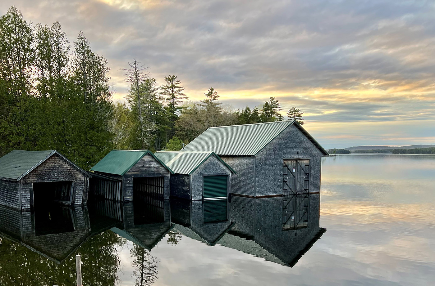 Boathouses on edge of water, with tall trees behind them 