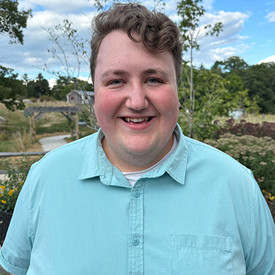 Alden-Hallett-1 Young man wearing light blue shirt, smiling in front of trees