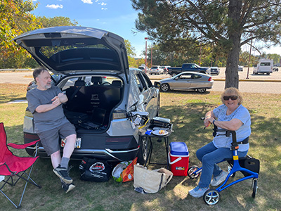 man sitting on open car trunk, woman sitting on walker to the right