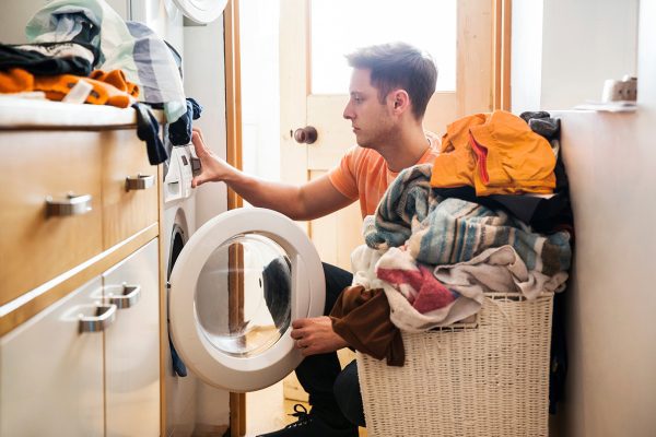 man kneeling down next to overflowing basket of laundry putting close in washing machine