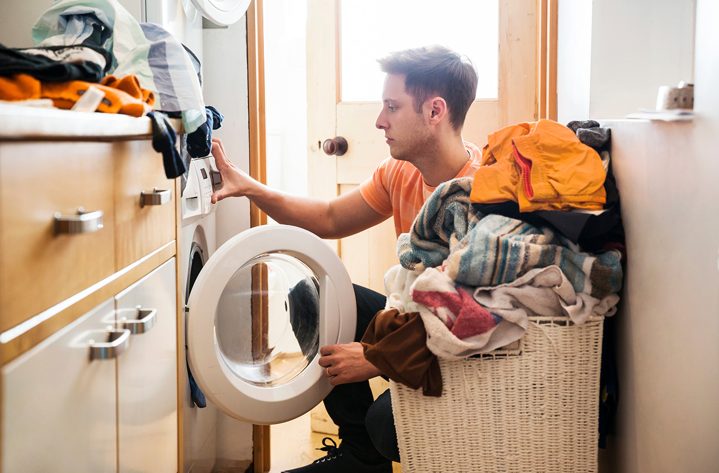 man kneeling down next to overflowing basket of laundry putting close in washing machine